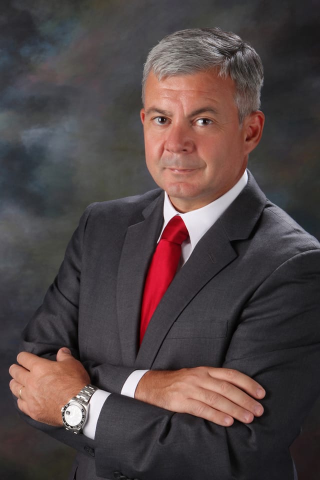 Retrato de Mario Panizza, Principal de Valmore Management & Staffing, con traje oscuro, camisa blanca y corbata roja sobre un fondo neutro suavemente difuminado.